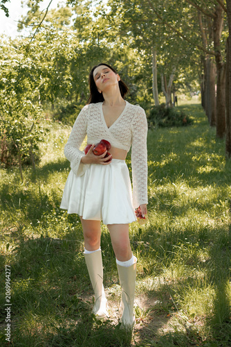 Creative portrait of woman in white lace outfit posing with red apples, balancing fruit on her head. Close up of girl with scraped, wounded knees covered with a green leaf in nature. Fairytale concept