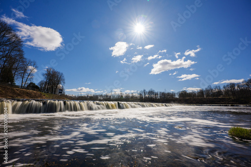 View of Ventas Rumba waterfall with flowing water under a bright sun and clear blue sky in Kuldiga, Kuldigas novads, Latvia.