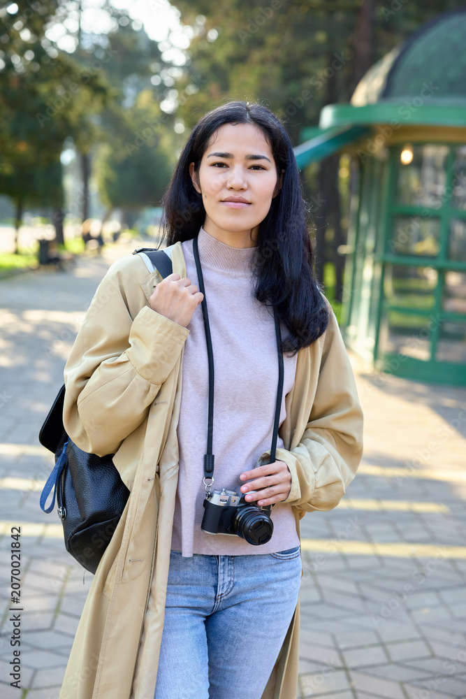 Fototapeta premium Woman with camera in an outdoor park setting, casual coat over sweater, backpack strap visible, street style photography and exploration vibe with natural light and calm ambience