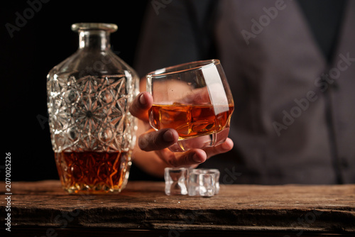 Man in Vest Holding Elegant Whiskey Glass with Crystal Decanter on Rustic Wooden Surface