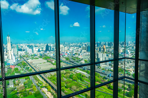 Aerial view of Bangkok, Thailand, seen through a glass window with dark framing, showing the skyline, rail lines, highways, and green urban spaces under a bright blue sky.