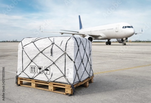 Wrapped pallet cargo sits on airport tarmac with blurred airplane in background representing global air freight logistics, shipment handling, and commercial aviation distribution operations industry