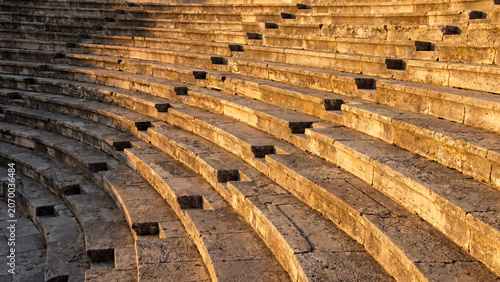 Ancient stone tiered seating rows of a historic mediterranean amphitheater glowing under the warm golden light of a late afternoon sun during a peaceful day at an archaeological ruin site.
