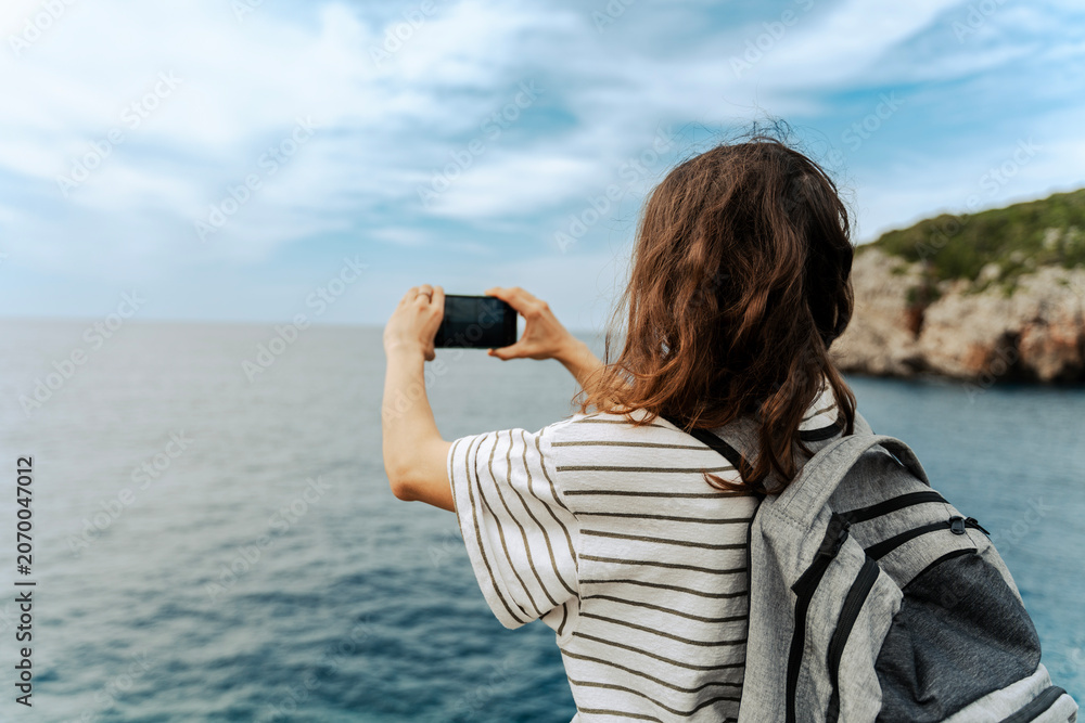 Naklejka premium Rear view of a female tourist with a backpack taking a photo of a scenic sea landscape with her smartphone during a journey