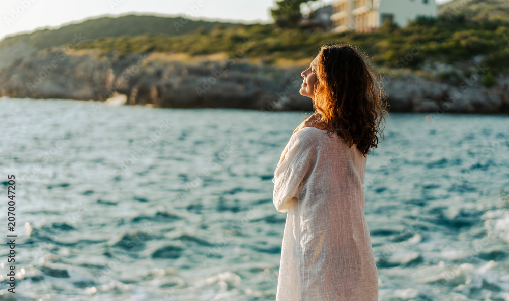 Obraz premium Side profile portrait of a beautiful young woman in a white shirt standing on the beach at sunset