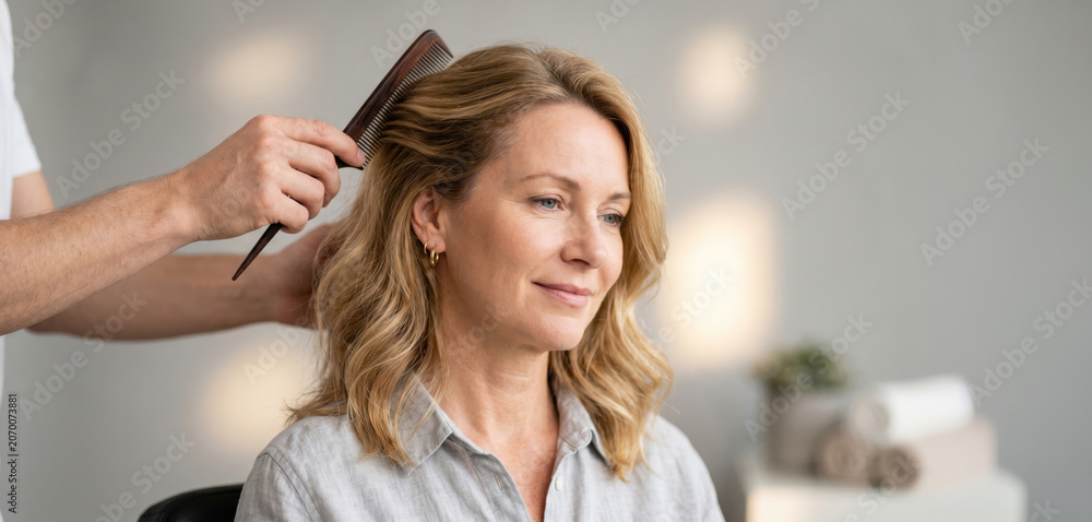 Naklejka premium A person is sitting in a salon chair while a stylist combs their hair. The salon has soft lighting. The stylist is focused on their work