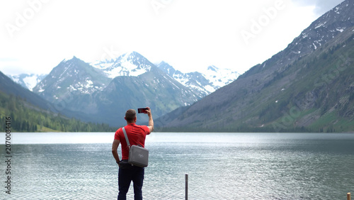 Photos An athletic male adventurer in a red t-shirt standing on a lake shore and using his smartphone to take a photo of distant snowy mountain peaks