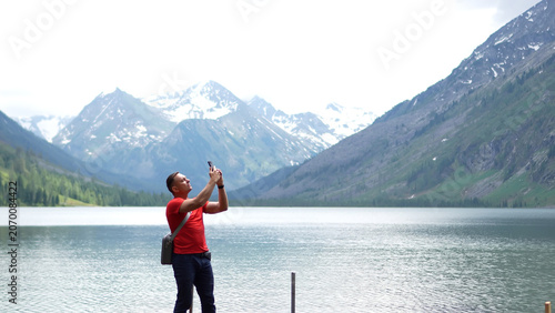 Obraz na plátně An athletic male adventurer in a red t-shirt standing on a lake shore and using his smartphone to take a photo of distant snowy mountain peaks