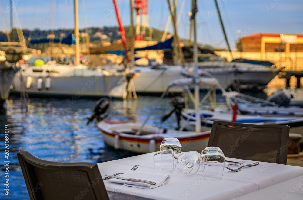 Fototapeta premium A table at an outdoor restaurant with a view of boats in the marina on the Mediterranean Sea in old town of Villefranche sur Mer, South of France