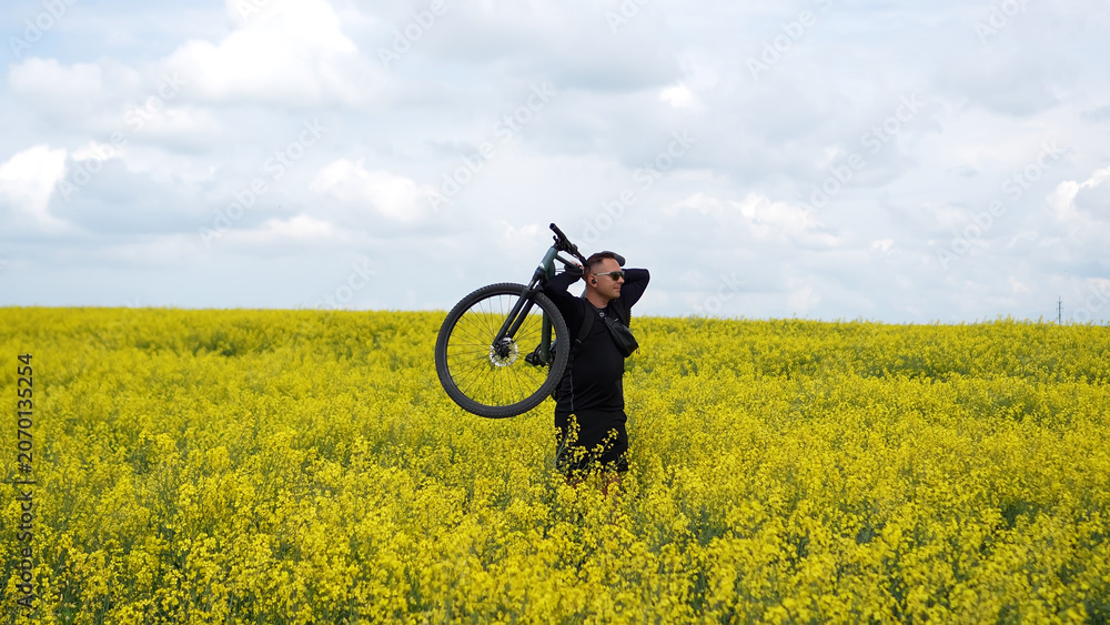 Fototapeta premium Moments of cycling. A man poses with his bicycle in a rapeseed field; a male athlete holds his bicycle behind his head, enjoying the views of the yellow rapeseed field.