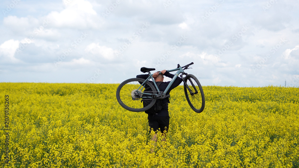 Fototapeta premium Moments of cycling. A man poses with his bicycle in a rapeseed field; a male athlete holds his bicycle behind his head, enjoying the views of the yellow rapeseed field.