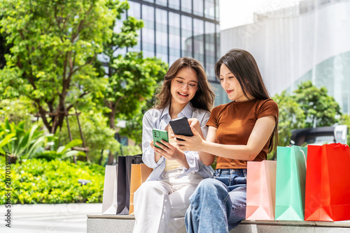 Two happy young women sit outdoors on a sunny day, holding smartphones and laughing while shopping. Vibrant colorful shopping bags surround them in a modern urban park setting, lifestyle photography.