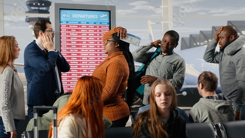 Photos Dejected by plane being late, FIDS screen and upset men with women in airport checking departure time, tedious waiting, electronic timetable display announcement and passengers bummed by flight delay