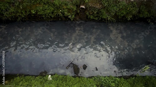Small Murky Water Stream Flowing Through Narrow Ditch With Green Vegetation On Both Sides