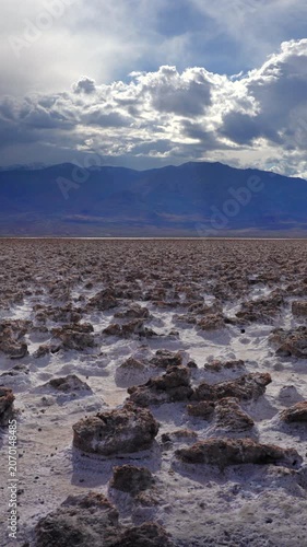Vertical Screen: Vast salt flat terrain with crystallized mineral formations stretches toward distant purple mountains under dramatic stormy clouds with sunlight breaking through.