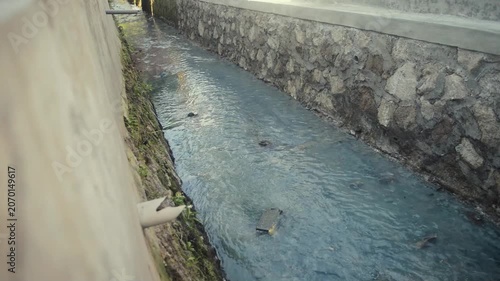 Polluted Gray Water Flowing Through Concrete Drainage Ditch With Trash Debris And Stone Walls