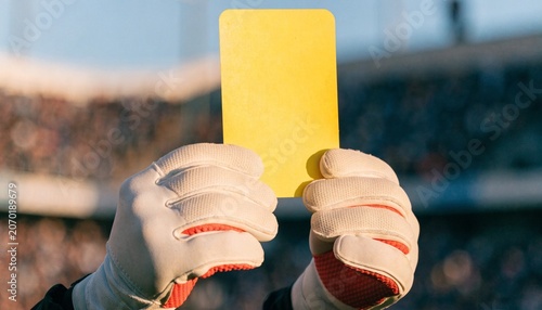 Close-up of a goalkeeper's hands in professional gloves holding up a bright blank yellow card against a blurred green football field background, indicating a penalty or warning during a match.