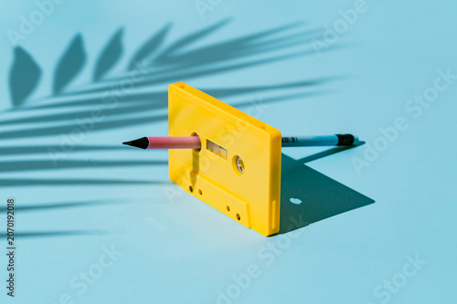 Tableau sur toile Creative retro summer music concept featuring a yellow audio cassette tape with a pencil inserted for manual rewinding and palm shadow on blue background