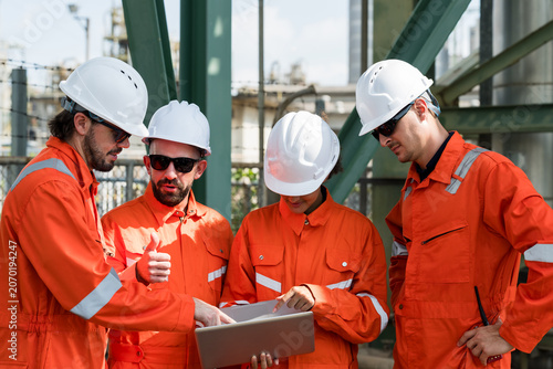 Multiracial refinery crew review laptop checklist during petrochemical maintenance briefing, coordinating pipeline safety inspection, compliance audit, operational risk assessment at industrial site