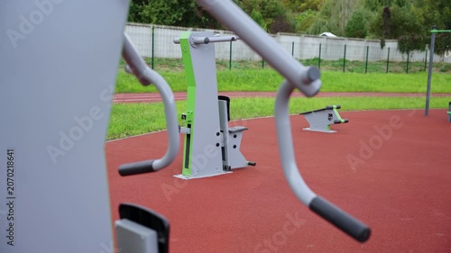 Outdoor gym equipment installed on red rubberized sports surface. Professional-grade machines situated near running track and lush lawn