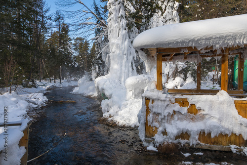 View of Goryachiye Istochniki Khakusy hot spring flowing through a snowy forest with a wooden shelter encased in ice in Severobaykal'skiy Rayon, Respublika Buryatiya, Russia.