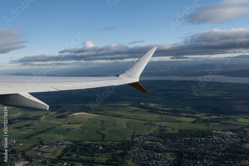 A scenic airplane window view showing the aircraft wing flying over the green fields, towns, and the Firth of Forth bay in Scotland