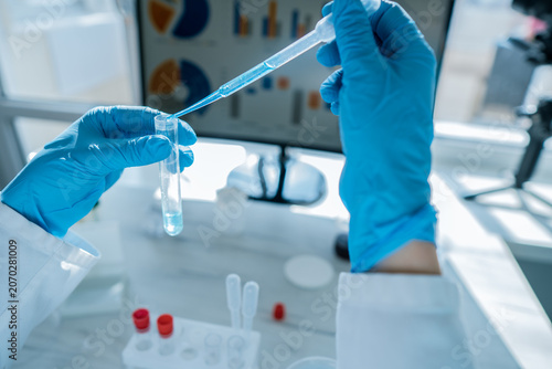 Close up of hands in protective gloves performing a laboratory experiment with a pipette and test tube. Advanced technology and scientific research tools are present on the workbench
