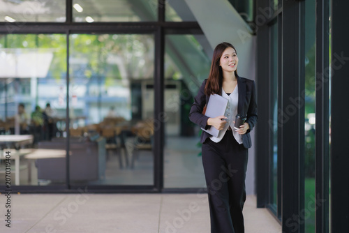 Young businesswoman walking through an office corridor, holding a laptop and a coffee cup, reflecting a professional and confident attitude in her daily routine