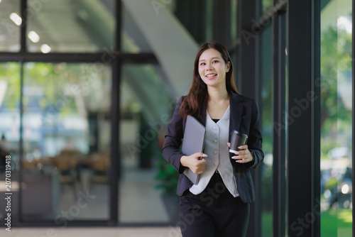 Young Asian businesswoman walking through a bright office hallway, carrying a laptop and coffee, symbolizing a dynamic professional lifestyle and entrepreneurial spirit