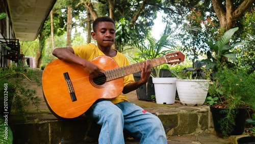 A stylish Indonesian teenager wearing a vibrant yellow t-shirt and blue jeans plays an acoustic guitar.