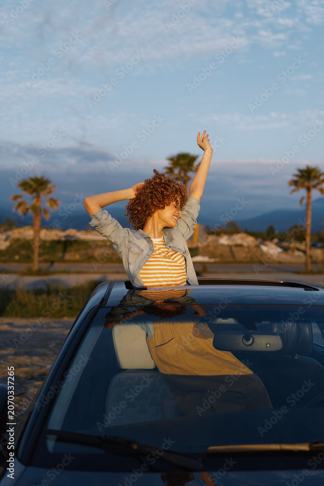 Fototapeta premium Young woman with curly hair leans out of a car window, arms raised in a carefree moment under a bright blue sky on a scenic roadside drive with warm sunlight and distant hills.