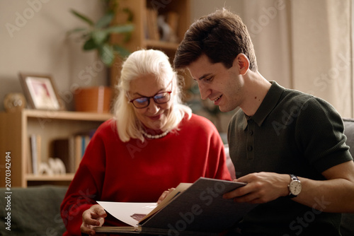 Fotografie Medium shot of senior Caucasian woman and young adult Caucasian man reviewing photo album at home, sharing family memories and bonding
