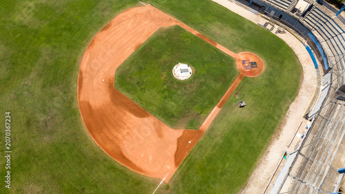 Photos Aerial drone shot of an empty baseball field diamond during a sunny morning, highlighting the red clay infield dirt, green grass turf, pitcher's mound, and clear white baseline markings clearly