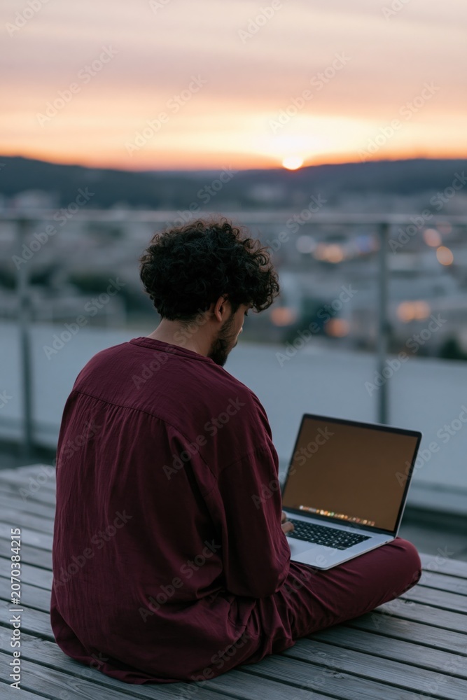 Obraz premium Man on rooftop working on laptop at sunset with city and hills in the distance for background and banner use