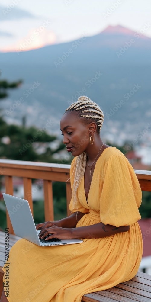 Fototapeta premium Young woman in yellow dress working on a laptop on a wooden balcony at sunset with mountain and city view for remote work background and lifestyle design