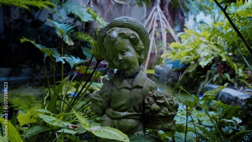 植物園の池のほとりに佇む苔むした子供の石像と緑豊かな庭園の風景 4Kシネマティック映像素材