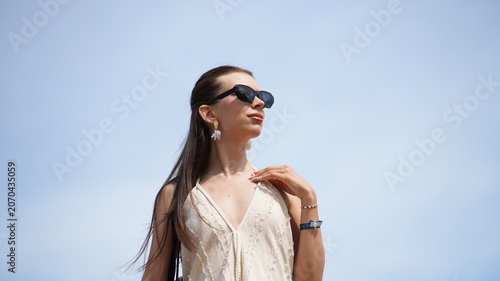 Schilderij op canvas Elegance Under the Sun: A stylish woman with long, dark hair and sunglasses poses confidently against a clear blue sky, embodying a sense of effortless chic and summer radiance
