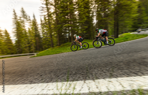 Two road cyclists riding downhill on a winding mountain road surrounded by forest. Dynamic motion blur captures speed, endurance training and outdoor cycling adventure.