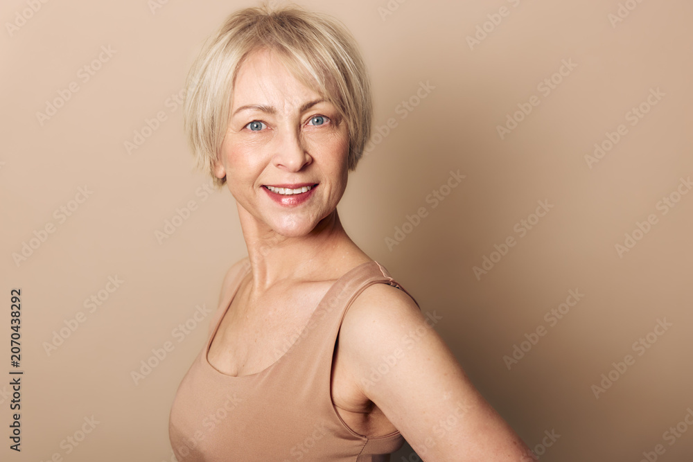 Fototapeta premium Smiling mature woman with short blonde hair wears beige tank top against neutral background. Studio portrait with soft lighting captures confident and natural beauty concept for aging gracefully.