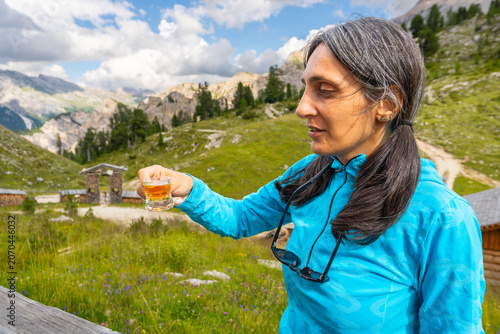 Schilderij op canvas Close up woman traveler drinking local herbal spirit schnapps or grappa shot glass wooden terrace green mountain valley backdrop Dolomites Italy