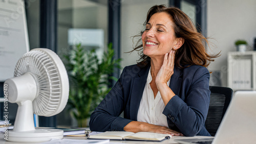 A woman in business attire enjoys a cool breeze from a desk fan while working at her laptop, smiling and looking refreshed in a modern office setting.