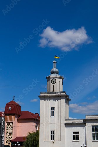 Historic town hall tower in Chortkiv, Ternopil region, Ukraine, featuring a clock and Ukrainian flag beneath a bright blue sky.