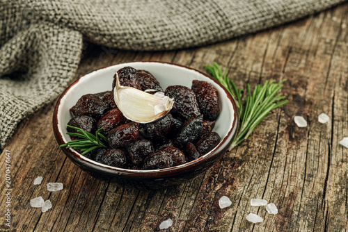 Black garlic cloves in oil with fresh rosemary branches and sea salt crystals in a ceramic bowl