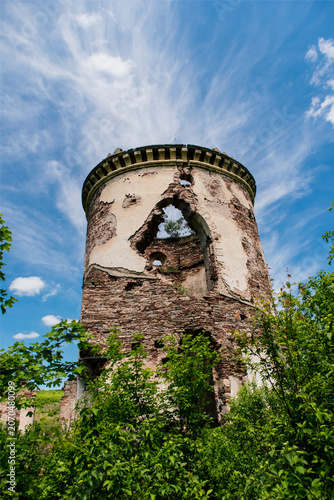 Weathered remains of the Chervonograd Castle tower stand among trees and shrubs, framed by bright clouds and clear blue skies.