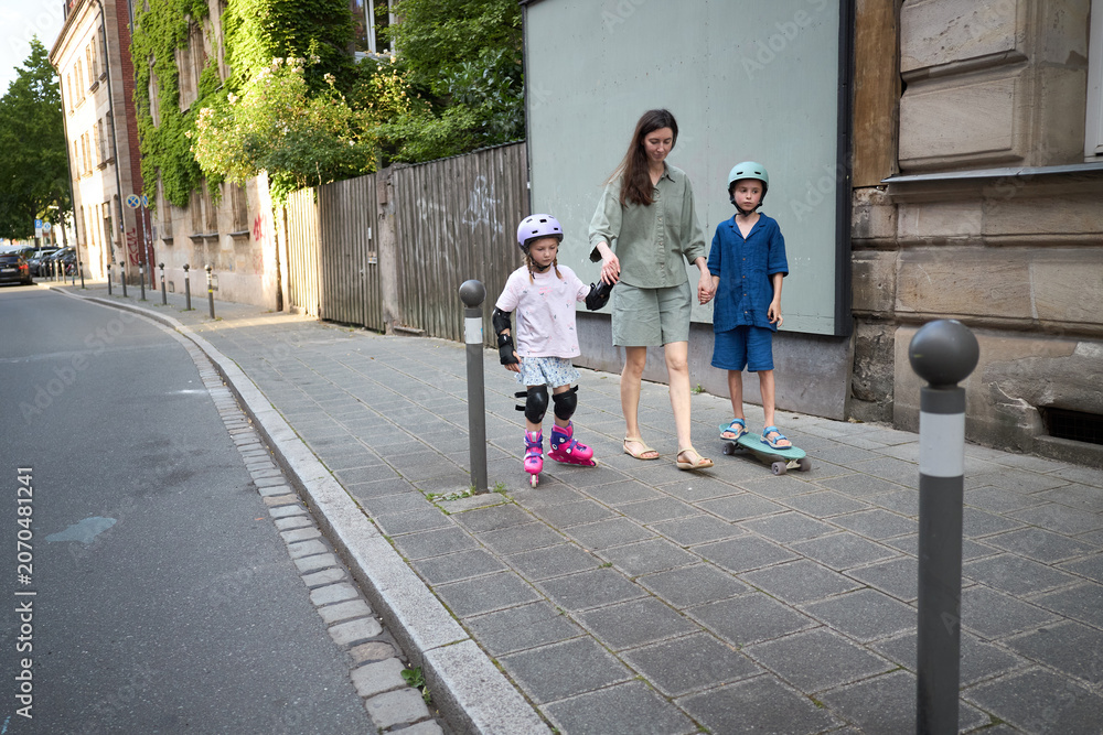 Fototapeta premium Mother supervises children rollerblading and skateboarding on a sunny city sidewalk