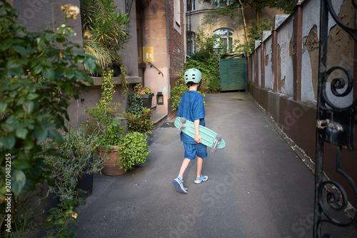 Young boy in helmet holding a skateboard walks down a narrow alleyway with plants
