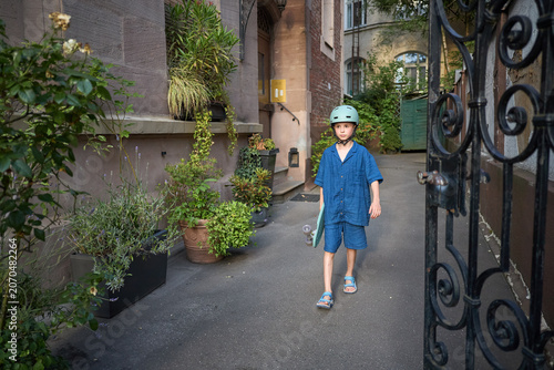 Young boy in blue outfit with skateboard walks through a gate in a courtyard