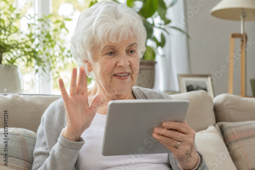 Grandmother sits on a comfortable living room sofa while waving to her family during a video call.