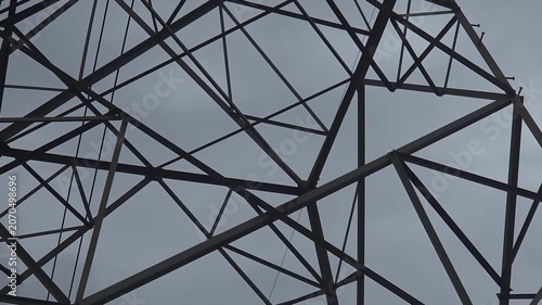 A high-voltage electricity pylon lattice frames a close-up view passing down to a hazard danger sign. Overcast clouds fill the background, signals power energy crisis, rain delays, weather uncertainty