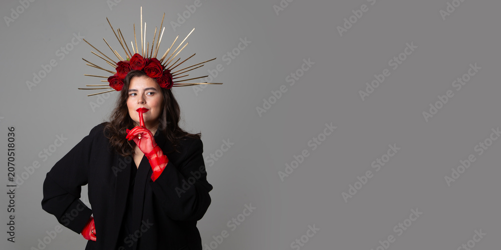 Obraz premium Photo banner. Young adult white woman with make up, black jacket, DIY golden halo crown, rose on head, red tulle gloves, choker, collar on neck raised index finger to lips on gray. Beauty. Copy space